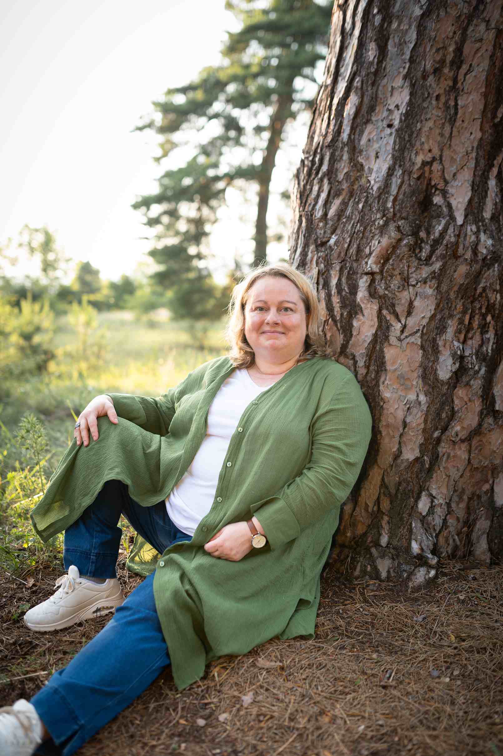Portrait von Eva Freyer sitzend vor einer Kiefer mit Natur im Hintergrund und Waldboden mit Nadeln