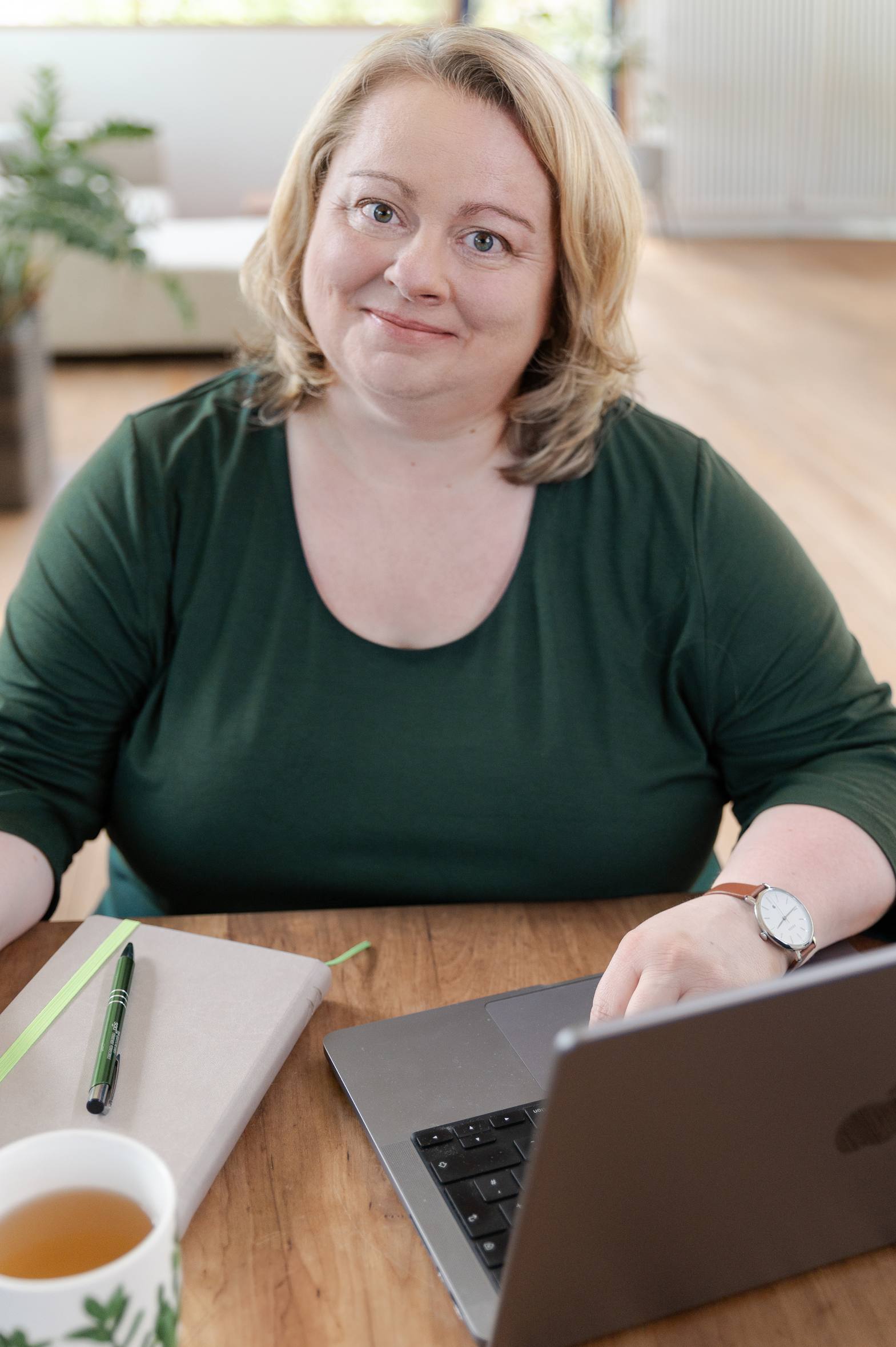 Portrait von Eva Freyer mit olivgrünem Oberteil am PC mit Notizbuch und Tasse vor hellem Hintergrund mit Holzboden