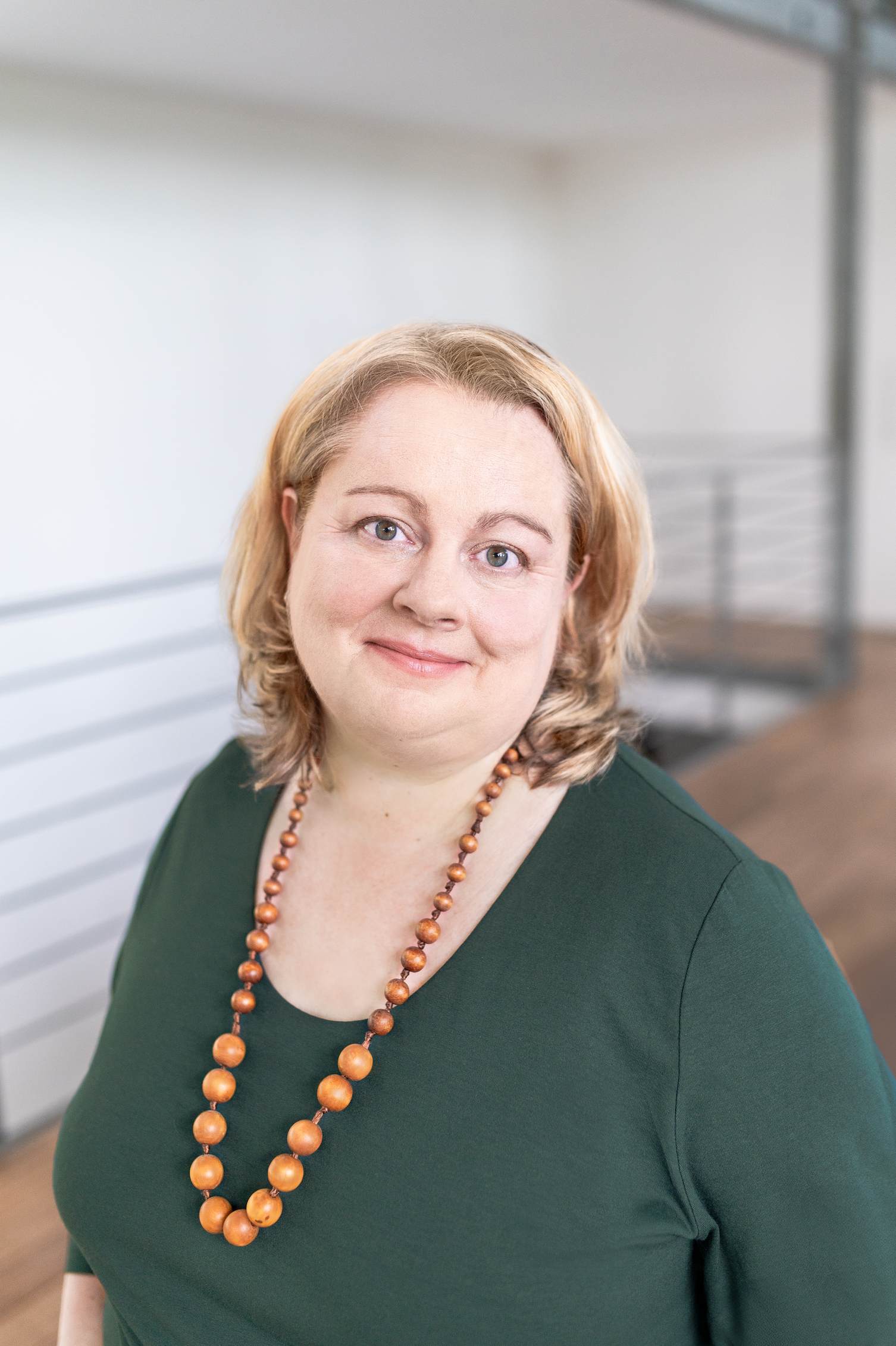 Portrait von Eva Freyer mit dunkelgrünen Oberteil und Holzkette vor hellem Hintergrund mit Holzboden