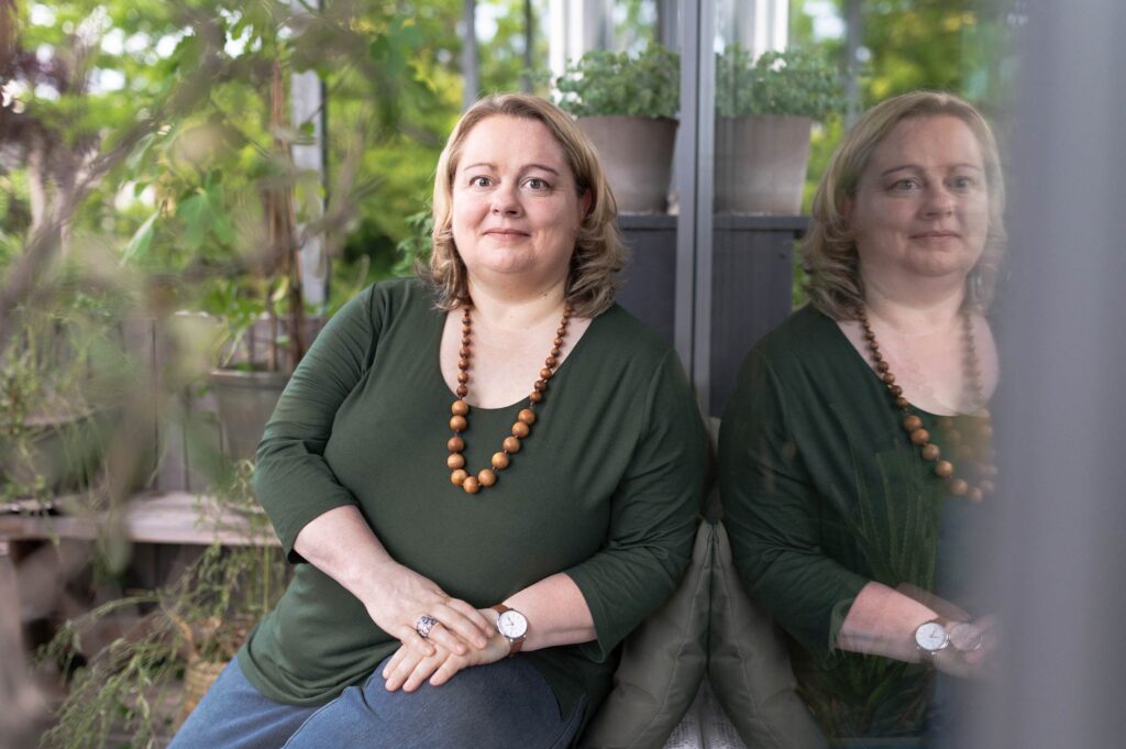 Portrait von Eva Freyer mit grünem Oberteil und Holzkette neben einer Glasschreib umgeben von grünen Pflanzen