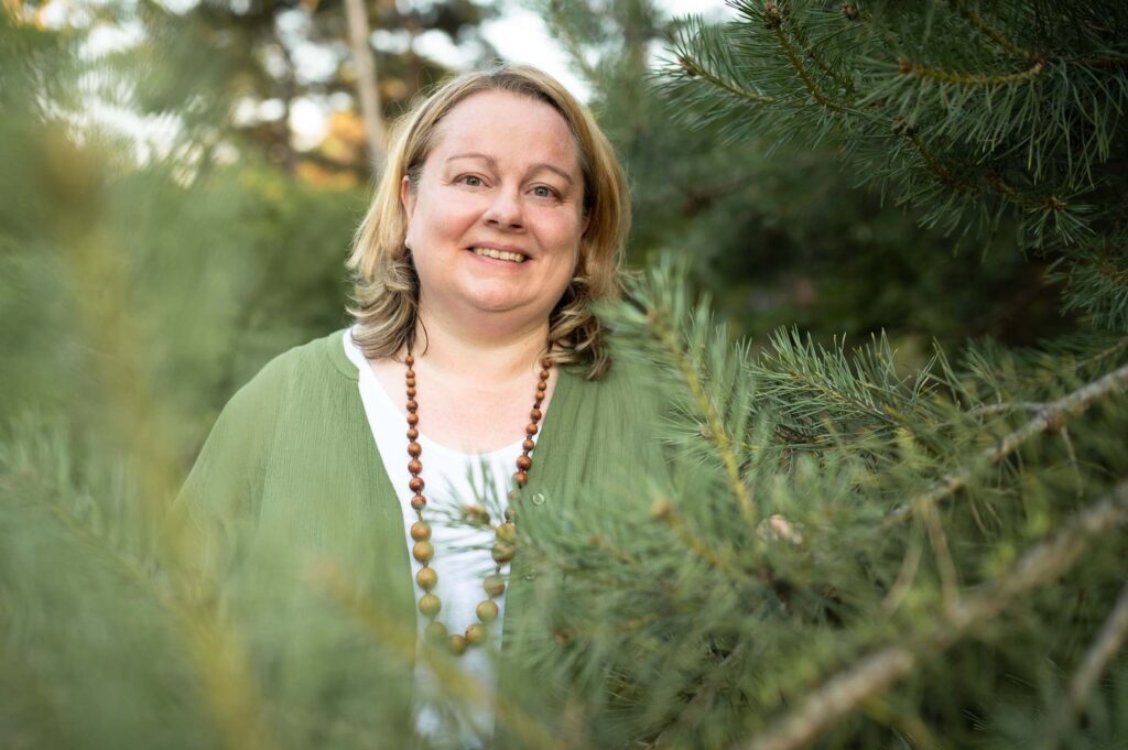 Portrait von Eva Freyer in grüner Bluse mit brauner Holzkette vor einer Kiefer mit grünem Hintergrund
