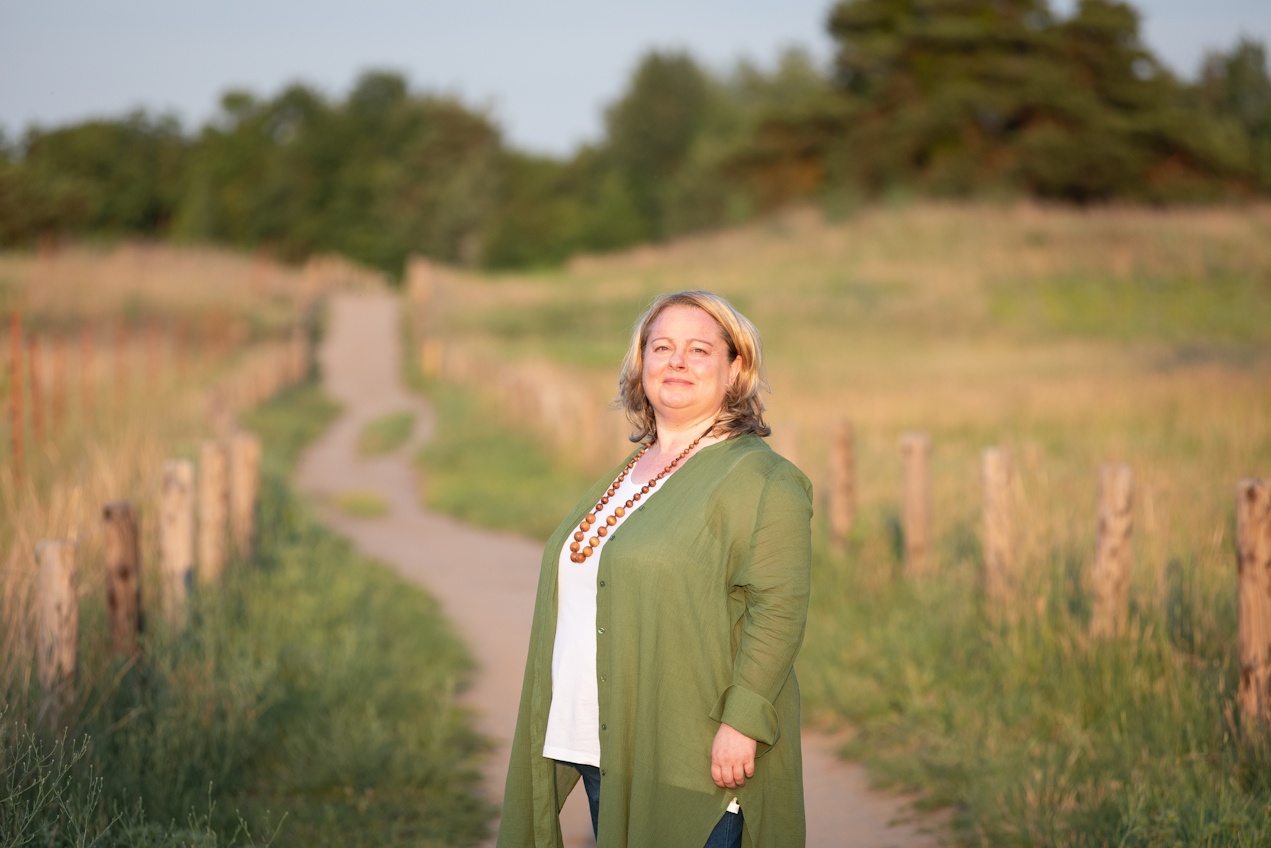Portrait von Eva Freyer in grüner Bluse mit Holzkette auf einem Sandweg mit Holzpfosten im Naturschutzgebiet mit grünen Sträuchern im Hintergrund und freier Wiese rechts und links des Wegs